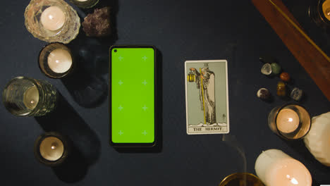 Draufsicht-Einer-Person,-Die-Neben-Der-Einsiedlerkarte-Auf-Dem-Tisch-Tarotkarten-Liest,-Mit-Einem-Mobiltelefon-Mit-Grünem-Bildschirm